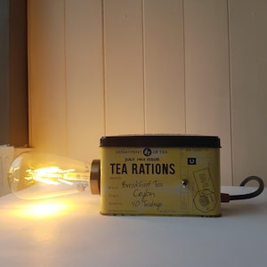 以下が含まれることがあります： ヴィンテージの紅茶缶で作られたユニークなランプ。 「TEA RATIONS」と書かれた黄色の缶には、黒い文字と黒い蓋があります。 エジソン風の電球が缶に取り付けられており、温かみのある光を放っています。 缶のテキストは「Breakfast Tea Ceylon」です。