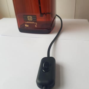 May include: A brown glass bottle with a black power cord and switch. The bottle has a label with the number 12 and other text. The switch is black and oval-shaped.