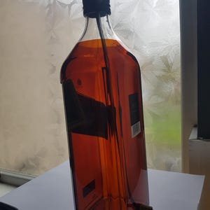 May include: A repurposed whiskey bottle converted into a lamp. The bottle is amber-colored with a black cap and a black electrical cord. The lamp is designed to be a unique home decor item.