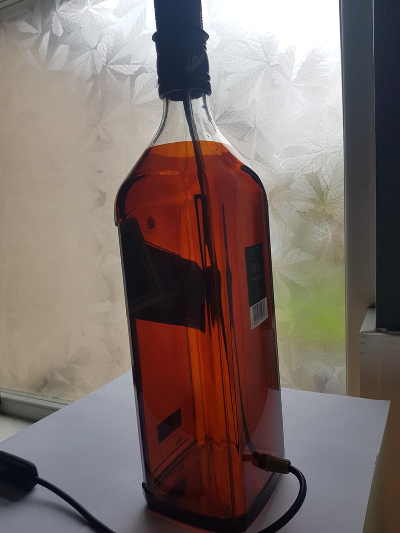 May include: A repurposed whiskey bottle converted into a lamp. The bottle is amber-colored with a black cap and a black electrical cord. The lamp is designed to be a unique home decor item.