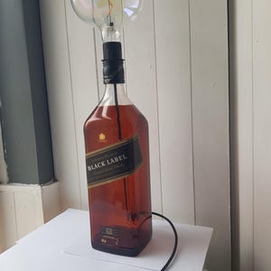 May include: A unique table lamp made from a Johnnie Walker Black Label whiskey bottle. The lamp features a large, round light bulb and a black cord with an on/off switch. The bottle is filled with a dark amber liquid.