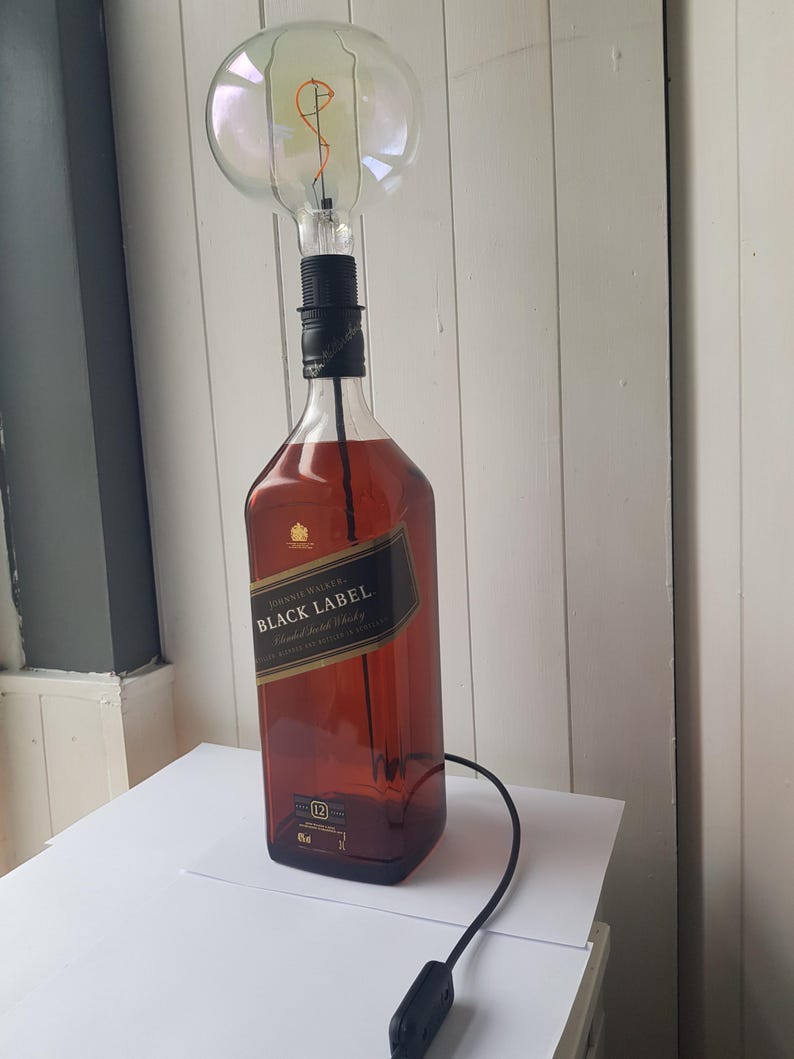 May include: A unique table lamp made from a Johnnie Walker Black Label whiskey bottle. The lamp features a large, round light bulb and a black cord with an on/off switch. The bottle is filled with a dark amber liquid.