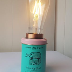 Könnte beinhalten: Eine einzigartige Tischlampe aus einer türkisfarbenen Fortnum & Mason Piccadilly Blend Kaffeedose. Die Lampe hat einen kupferfarbenen Sockel, einen rosa Rand und eine große Edison-Glühbirne. Die Dose zeigt eine Pelikan-Illustration und den Text "Piccadilly Blend".