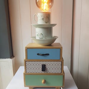 May include: A table lamp featuring a stack of white floral ceramic cups and bowls topped with a glowing Edison bulb, resting on a wooden three-drawer chest with blue, patterned, and green drawer fronts and a white power cord with a switch.