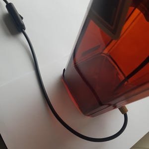 May include: A rectangular amber-colored lamp with a black power cord and switch. The lamp has a geometric design and a brass-colored fitting where the cord connects. The lamp is on a white surface.