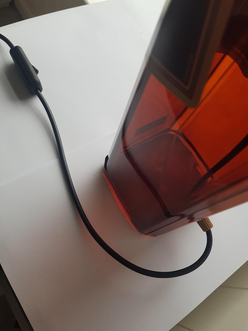 May include: A rectangular amber-colored lamp with a black power cord and switch. The lamp has a geometric design and a brass-colored fitting where the cord connects. The lamp is on a white surface.