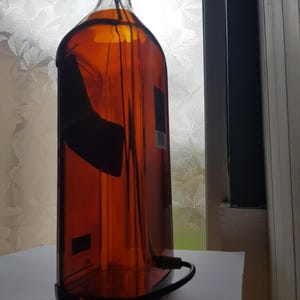 May include: A repurposed amber glass bottle with a black cap and a black power cord. The bottle has a rectangular shape and a dark label. The bottle is sitting on a white surface.