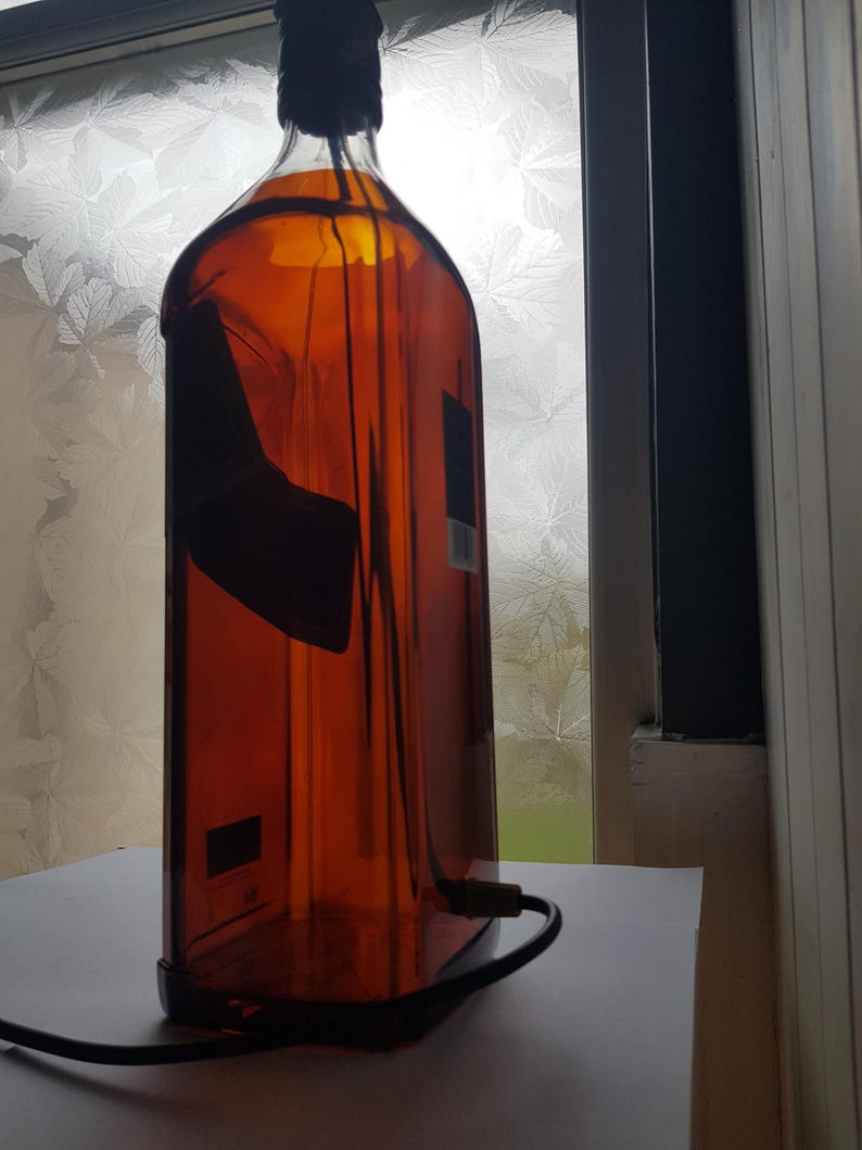 May include: A repurposed amber glass bottle with a black cap and a black power cord. The bottle has a rectangular shape and a dark label. The bottle is sitting on a white surface.