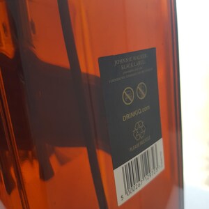 May include: A close-up of a brown glass bottle with a black label. The label reads "Johnnie Walker Black Label" and includes the website "DrinkIQ.com". The bottle has a barcode and a recycling symbol.