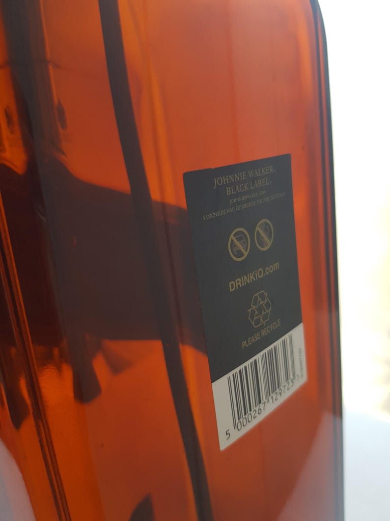 May include: A close-up of a brown glass bottle with a black label. The label reads "Johnnie Walker Black Label" and includes the website "DrinkIQ.com". The bottle has a barcode and a recycling symbol.