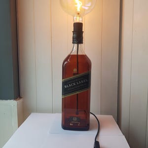 May include: A unique table lamp made from a repurposed Johnnie Walker Black Label whiskey bottle. The lamp features a large, round Edison-style light bulb and a black cord with an on/off switch. The bottle is brown with a black and gold label.