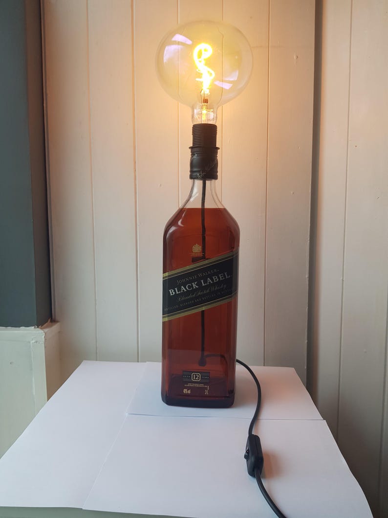 May include: A unique table lamp made from a repurposed Johnnie Walker Black Label whiskey bottle. The lamp features a large, round Edison-style light bulb and a black cord with an on/off switch. The bottle is brown with a black and gold label.