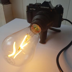 Könnte beinhalten: Eine schwarze Cosina CT1G Filmkamera, die als Lampe umfunktioniert wurde, mit einer großen, klaren Edison-Glühbirne aus Glas, die aus dem Objektivanschluss ragt. Das Kameragehäuse ist schwarz mit weißer Schrift und hat ein schwarzes Stromkabel.