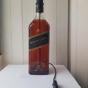 May include: A repurposed Johnnie Walker Black Label whiskey bottle transformed into a lamp. The amber-colored glass bottle features a black label with gold lettering. A black lamp socket and cord with an on/off switch are attached.