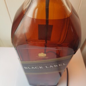 May include: A partially filled bottle of Johnnie Walker Black Label whiskey. The amber liquid is visible through the clear glass. The label is black and gold, with the brand name prominently displayed. A black cord extends from the bottle.