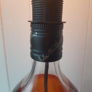 May include: A repurposed liquor bottle lamp with a black lamp holder and a clear lightbulb. The bottle is filled with a brown liquid and has the words "John Walker" in cursive script on the black cap.