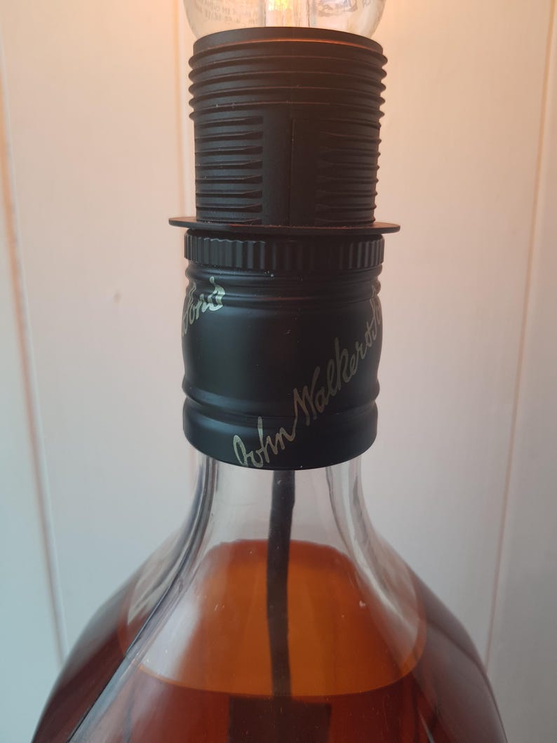 May include: A repurposed liquor bottle lamp with a black lamp holder and a clear lightbulb. The bottle is filled with a brown liquid and has the words "John Walker" in cursive script on the black cap.