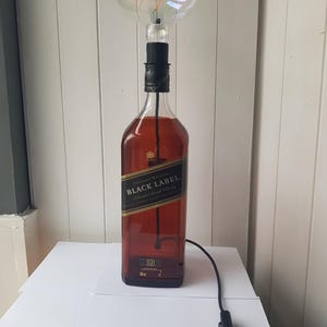 May include: A unique lamp made from a Johnnie Walker Black Label bottle. The lamp features a large, round light bulb and a black cord with an on/off switch. The bottle has a black and gold label.