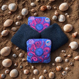 May include: Four bean bags with a dark blue fabric and two with a vibrant octopus design in pink and blue. The bags are surrounded by seashells on a sandy surface. The bags have the text "D8" and a logo.