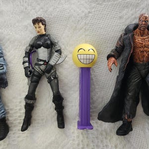 May include: Four action figures, including a man in a blue jumpsuit, a woman in a black and silver outfit, a man in a brown coat with a bare chest, and a yellow smiley face Pez dispenser.