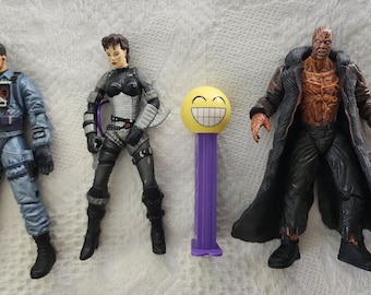 May include: Four action figures, including a man in a blue jumpsuit, a woman in a black and silver outfit, a man in a brown coat with a bare chest, and a yellow smiley face Pez dispenser.