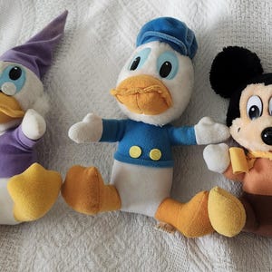 Puede incluir: Tres peluches de personajes de Disney: Pato Donald con camisa y gorro de marinero azules, un pato morado y Mickey Mouse en marr&oacute;n y naranja. Los juguetes est&aacute;n sobre una superficie blanca.