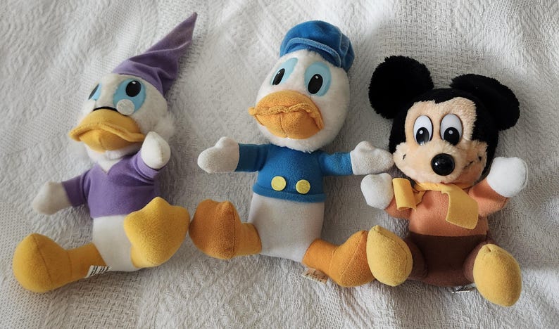 Puede incluir: Tres peluches de personajes de Disney: Pato Donald con camisa y gorro de marinero azules, un pato morado y Mickey Mouse en marr&oacute;n y naranja. Los juguetes est&aacute;n sobre una superficie blanca.