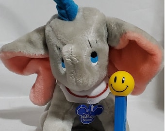 May include: A gray plush Dumbo the elephant toy with a blue hat and pink ears. Dumbo is wearing a red and white collar with a blue Disney original tag. A blue and yellow smiley face lollipop is next to Dumbo.