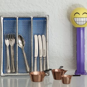 May include: A silver cutlery set with forks, spoons, and knives in a blue mesh holder. Three small copper pots with black handles are in front of the cutlery. A yellow smiley face Pez dispenser with a purple base is on the right.