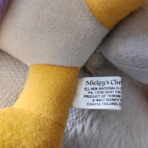 Puede incluir: Primer plano de un peluche con cuello amarillo y etiqueta blanca. La etiqueta dice "Mickey's Chr" e incluye texto sobre materiales y origen. El juguete tiene un cuerpo gris y una manga morada.