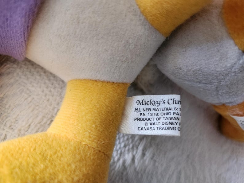 Puede incluir: Primer plano de un peluche con cuello amarillo y etiqueta blanca. La etiqueta dice "Mickey's Chr" e incluye texto sobre materiales y origen. El juguete tiene un cuerpo gris y una manga morada.