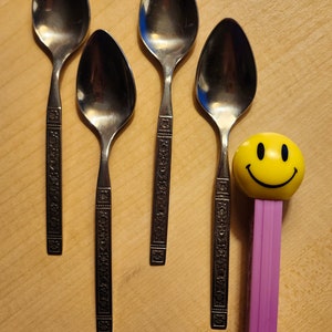 Pode incluir: Quatro colheres de prata com desenhos ornamentados nas alças, dispostas em fila sobre uma superfície de madeira. Um brinquedo de cara sorridente roxo está à direita das colheres.