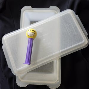 May include: A clear plastic rectangular container with a lid, and a yellow and purple Pez dispenser with a smiling face. The container is labeled "UPPER".