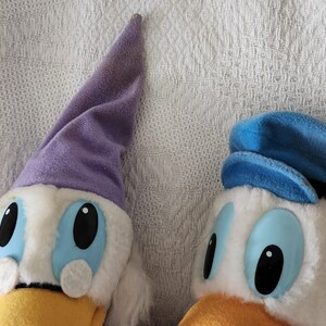 Puede incluir: Dos peluches de Pato Donald. Uno lleva un gorro de dormir morado, el otro un sombrero de marinero azul. Ambos tienen pelaje blanco, ojos azules y picos amarillos. Los juguetes est&aacute;n sobre un fondo blanco texturizado.