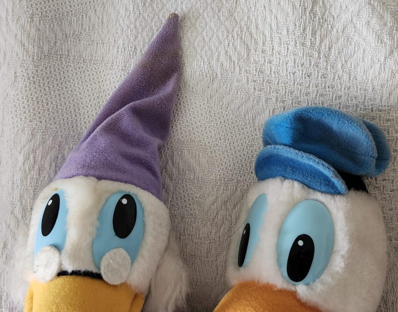 Puede incluir: Dos peluches de Pato Donald. Uno lleva un gorro de dormir morado, el otro un sombrero de marinero azul. Ambos tienen pelaje blanco, ojos azules y picos amarillos. Los juguetes est&aacute;n sobre un fondo blanco texturizado.