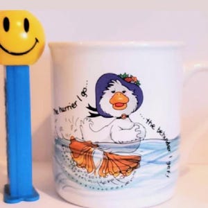 May include: A yellow smiley face PEZ dispenser next to a white ceramic mug. The mug features a cartoon duck in a blue hat and the text "...the hurrier I go...the behinder I get!"