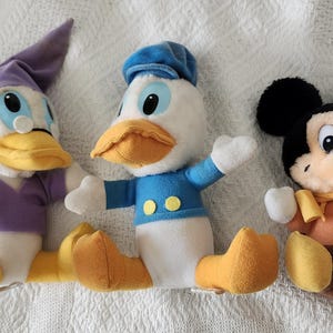 Puede incluir: Tres peluches de personajes de Disney: Pato Donald, Mickey Mouse y un personaje parecido a un pato. Pato Donald viste una camisa y gorro de marinero azules. Mickey Mouse tiene orejas negras y cuerpo marr&oacute;n. El personaje parecido a un pato lleva gorro y camisa morados.
