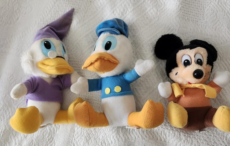 Puede incluir: Tres peluches de personajes de Disney: Pato Donald, Mickey Mouse y un personaje parecido a un pato. Pato Donald viste una camisa y gorro de marinero azules. Mickey Mouse tiene orejas negras y cuerpo marr&oacute;n. El personaje parecido a un pato lleva gorro y camisa morados.