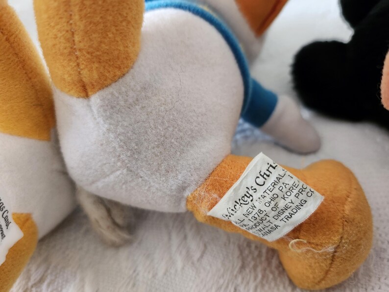Puede incluir: Primer plano de un peluche de Donald Duck. El juguete tiene un cuerpo blanco, piernas naranjas y una camisa azul. Una etiqueta en la pierna dice "Mickey's Christmas". El juguete est&aacute; sobre una superficie blanca.