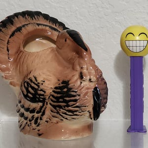 May include: A ceramic turkey-shaped container with brown, black, and tan details. Next to it is a yellow smiley face PEZ dispenser with a purple base. The turkey has a detailed feather design and a hollowed-out center.