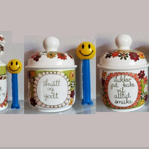 May include: Three ceramic sugar jars with floral designs and lids. The jars have different sayings on them: "Socker är sött men tål ej blött", "Smått og godt", and "Sukker på kake vil alltid smake". Each jar is next to a blue and yellow smiley face lollipop.