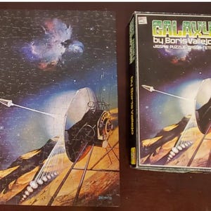 Puede incluir: Un rompecabezas completado y su caja de cartón rectangular con la obra de arte Galaxy de Boris Vallejo. La imagen muestra una gran antena parabólica sobre la superficie rocosa y anaranjada de un planeta con un fondo espacial azul oscuro.