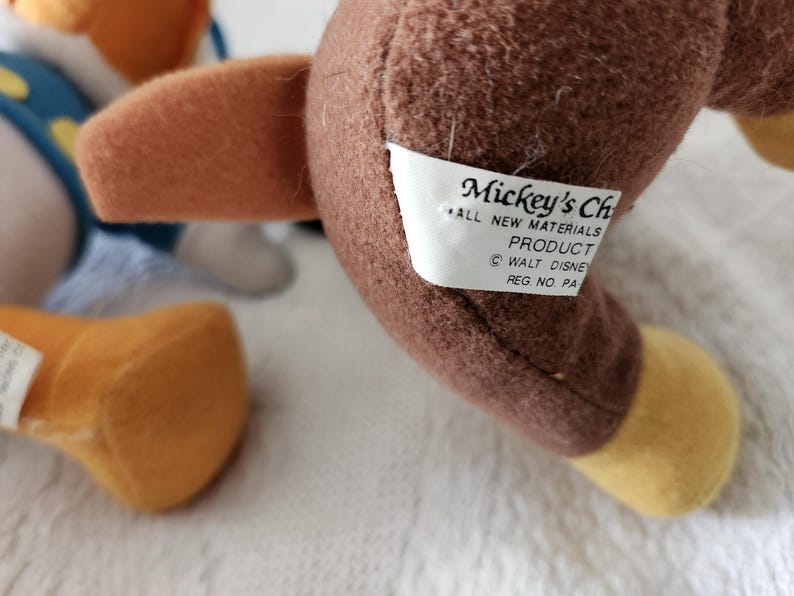 Puede incluir: Primer plano de un juguete de peluche marr&oacute;n con un ala marr&oacute;n y un pie amarillo. Una etiqueta blanca dice "Mickey's Ch... ALL NEW MATERIALS PRODUCT &copy; WALT DISNEY REG. NO. PA".