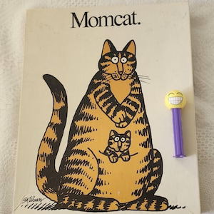 Puede incluir: Una portada de libro con el título "Momcat" y una gran ilustración de gato de dibujos animados en naranja y negro. Un gato pequeño está en la bolsa del gato más grande. A la derecha hay un dispensador de Pez amarillo y morado con una cara sonriente.