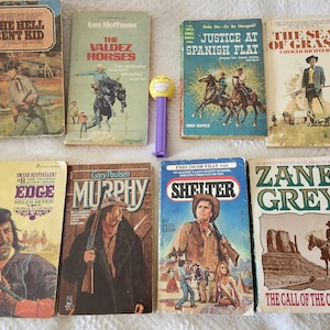 May include: Collection of vintage paperback books with Western themes. Titles include "The Hell Bent Kid," "The Valdez Horses," "Justice at Spanish Flat," and "The Sea of Grass." A Pez dispenser is also visible.