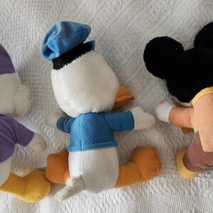 Puede incluir: Tres peluches de personajes de Disney: Pato Donald, Mickey Mouse y un tercer personaje. El Pato Donald lleva una camisa y un sombrero azules. Mickey Mouse tiene orejas negras y un atuendo marr&oacute;n. El tercer personaje lleva un atuendo morado.