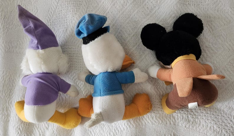 Puede incluir: Tres peluches de personajes de Disney: Pato Donald, Mickey Mouse y un tercer personaje. El Pato Donald lleva una camisa y un sombrero azules. Mickey Mouse tiene orejas negras y un atuendo marr&oacute;n. El tercer personaje lleva un atuendo morado.