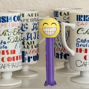 Könnte beinhalten: Weiße Keramikbecher mit Sockel und gebogenem Griff, mit Text wie "Irish Coffee" und "Cafe Brule". Ein gelber Pez-Spender mit Smiley-Gesicht und lila Basis befindet sich vor den Bechern.