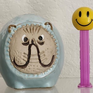 Może przedstawiać: Ceramiczna skarbonka w kształcie buldoga w kolorze jasnoniebieskim i beżowym ze szczegółową twarzą. Obok żółty dozownik PEZ z uśmiechniętą buzią i różowym patyczkiem. Przedmioty znajdują się na odblaskowej powierzchni.