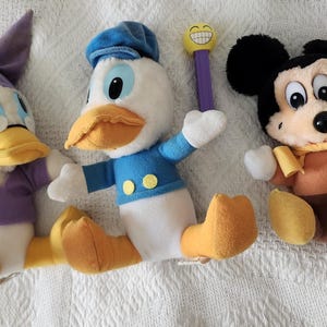 Puede incluir: Tres peluches de personajes de Disney: Pato Donald, Mickey Mouse y una variante de Pato Donald. Pato Donald viste camisa y gorro de marinero azules, Mickey Mouse tiene cuerpo marr&oacute;n y orejas negras. La variante de Pato Donald viste un atuendo morado.